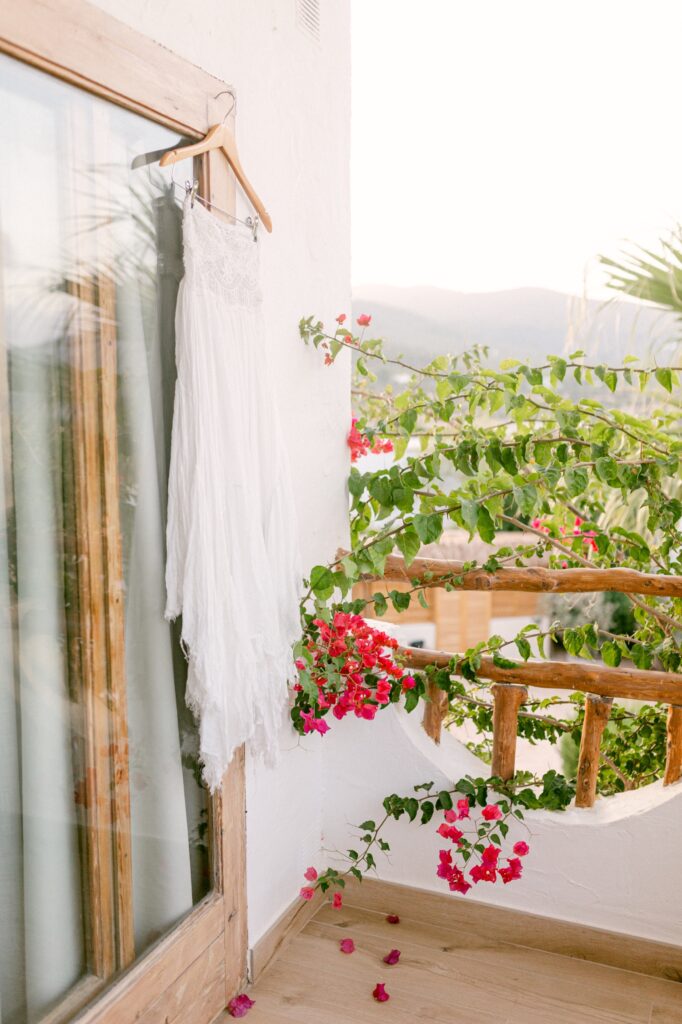 An elegant white wedding dress hanging by vibrant bougainvillea flowers at a picturesque Ibiza villa, glowing softly in the warm hues of sunrise, captured by Jenni Summer Studios.