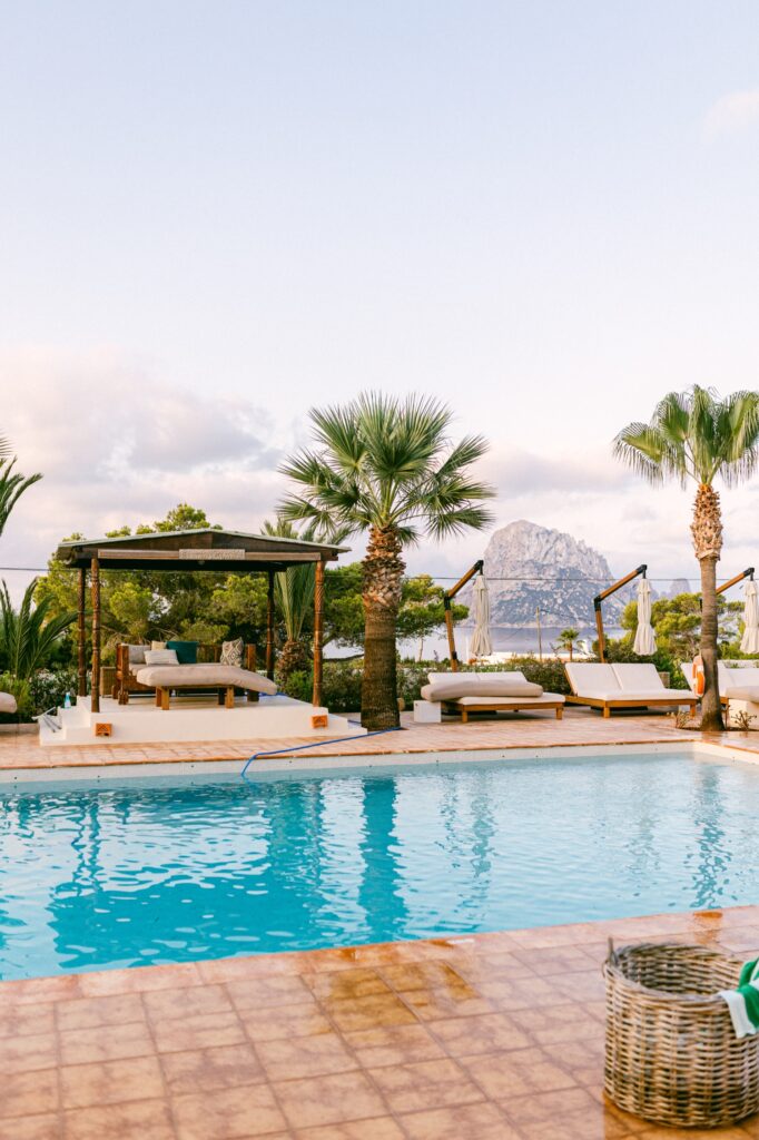 The serene pool at Petunia Ibiza, a Beaumier Hotel, with elegant cabanas lining the edge and the stunning Es Vedra rock formation in the background, bathed in the soft light of sunrise, captured by Jenni Summer Studios