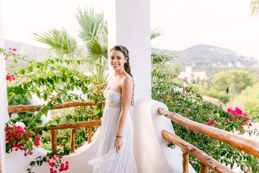 The bride standing gracefully on the balcony in her sleeveless bohemian-style wedding dress, with vibrant bougainvillea flowers framing the background, captured in a romantic moment during her Ibiza elopement by Jenni Summer Studios